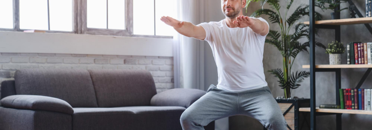 Een man staat op een matje in een woonkamer. Hij traint met zijn eigen lichaamsgewicht. Hij heeft zijn knieën gebogen en strekt zijn armen naar voren.