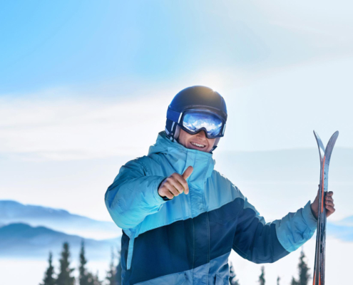 Een man staat in de sneeuw en heeft in zijn ene hand ski's vast en met zijn andere hand steekt hij zijn skiduim op.