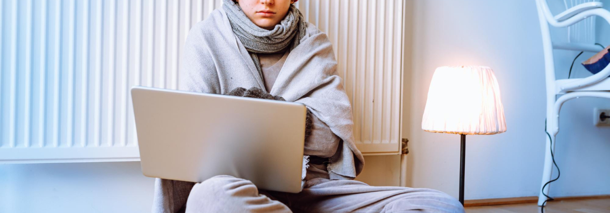 Persoon zit somber tegen verwarming aan op de grond met muts op en laptop op schoot. Persoon heeft herfst- en wintermoeheid.