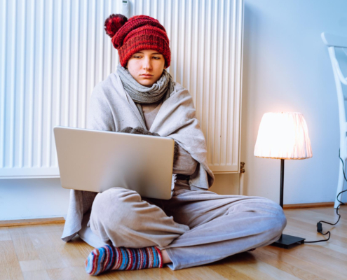 Persoon zit somber tegen verwarming aan op de grond met muts op en laptop op schoot. Persoon heeft herfst- en wintermoeheid.