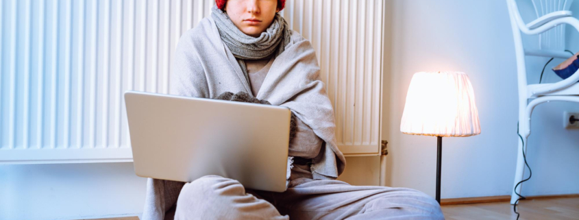 Persoon zit somber tegen verwarming aan op de grond met muts op en laptop op schoot. Persoon heeft herfst- en wintermoeheid.