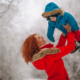 Vrouw in een rode jas heeft geen last van Rugpijn in de winter en tilt haar kind in een blauwe jas op.