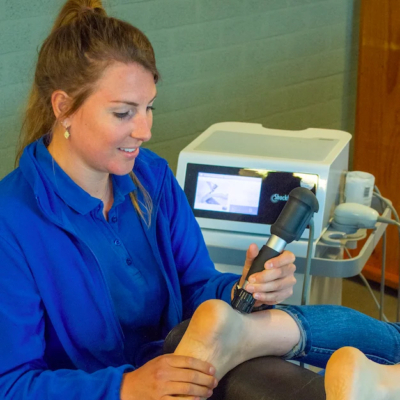 Shockwave therapie door een vrouwelijke fysiotherapeut in een blauw shirt bij de hiel van een patiënt
